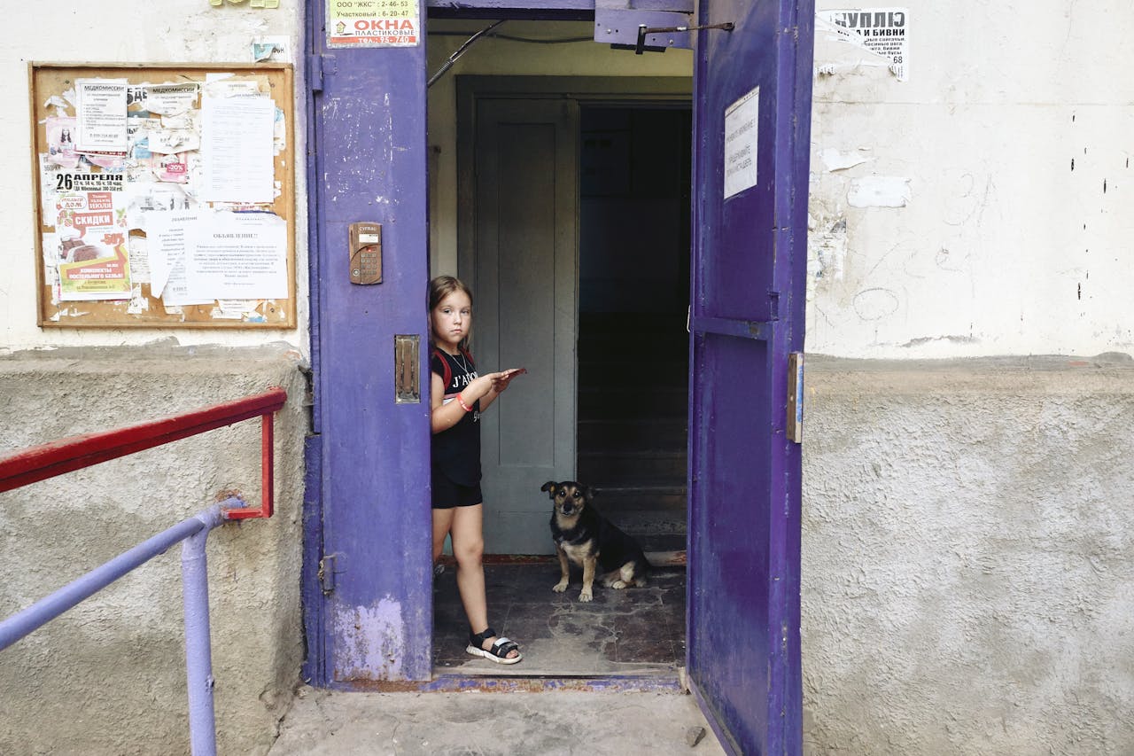 A young girl and her dog stand in a purple doorway of an urban building.